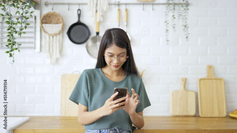 Happy young asian woman wearing glasses and relaxing at home. Asia female standing at counter kitchen and using mobile smartphone Happy young asian woman wearing glasses and relaxing at home. Asia female standing at counter kitchen and using mobile smartphone