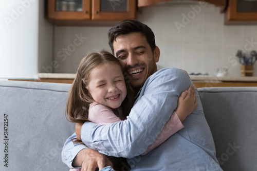 Adorable preschool daughter cuddles tightly her stepfather with eyes closed seated on sofa at home, enjoy tender moment, reunion after long separation, feels love, express caress. Family ties concept