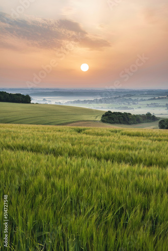 Beautiful English Summer sunrise landscape image looking over rolling hills with mist in distance and hazy sun low in the sky