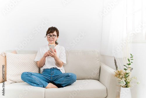 家のソファでスマホを使うメガネの女性（笑顔）
