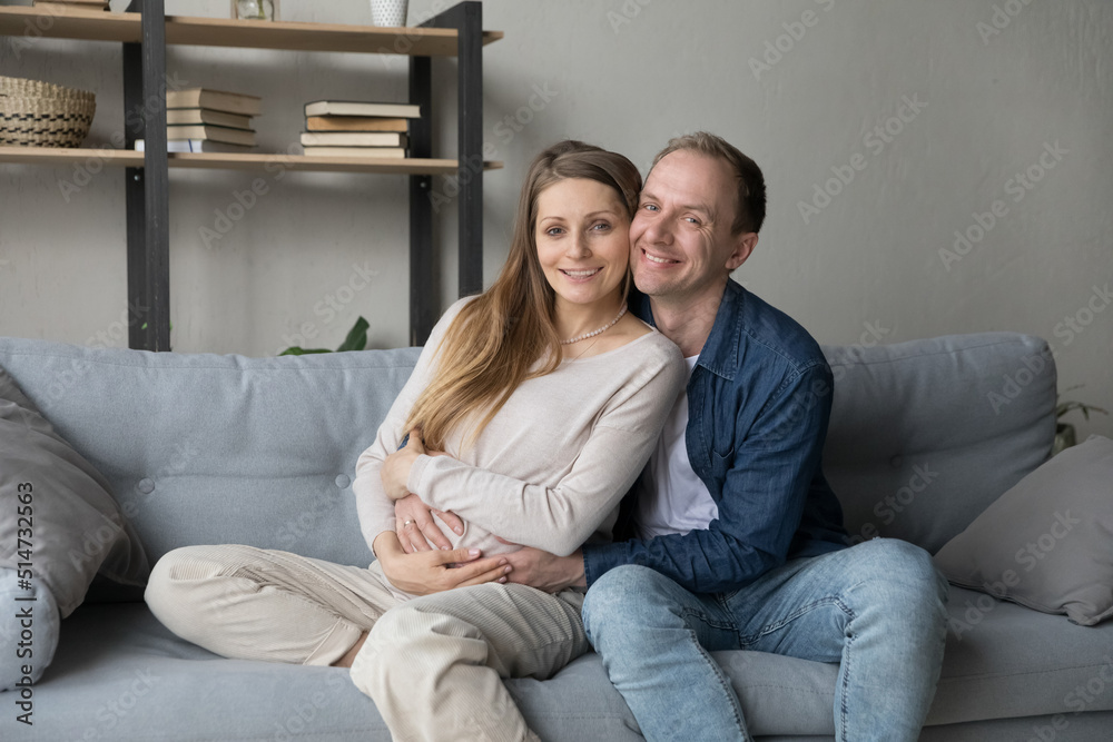 Young 30s attractive spouses sitting on sofa posing for family portrait, pregnant woman her caring husband touch belly feeling happy spend time together at home. Bank loan, first time parents concept Young 30s attractive spouses sitting on sofa posing for family portrait, pregnant woman her caring husband touch belly feeling happy spend time together at home. Bank loan, first time parents concept