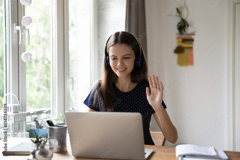 Happy student woman in wireless headphones talking to teacher on video call, attending online class, conference. Blogger girl greeting audience, waving hand hello at laptop webcam