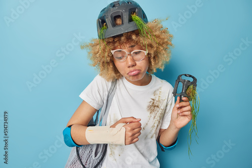 Unhappy curly woman wears bandage on broken arm after dangerous bicycle riding wears protective helmet and elbow pads dressed in white t shirt holds pedal from bike isolated over blue background