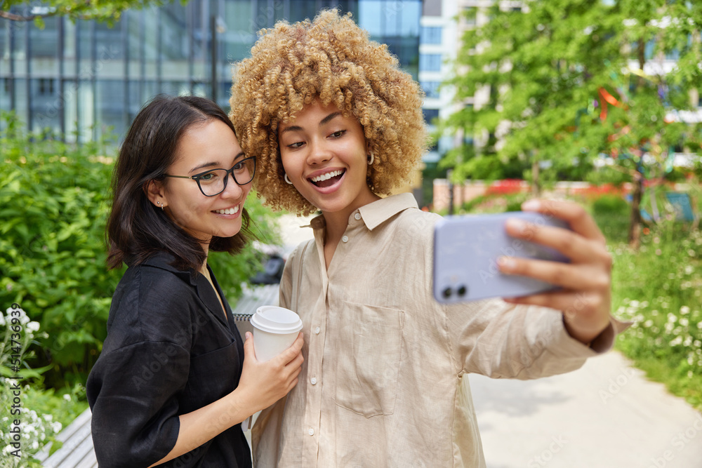 Multiracial women friends spends free time together in city pose for making selfie drink takeaway coffee wear casual clothes have good mood. Female bloggers talk to followers record streaming video