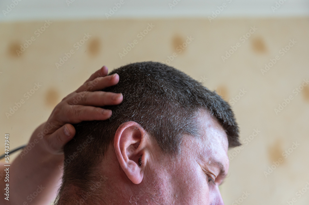 Obraz premium A woman cuts a man's hair. The persona sits against a yellow wall. The woman's hand rests on the back of the man's head. Short brown hair. Side view. Selective focus.