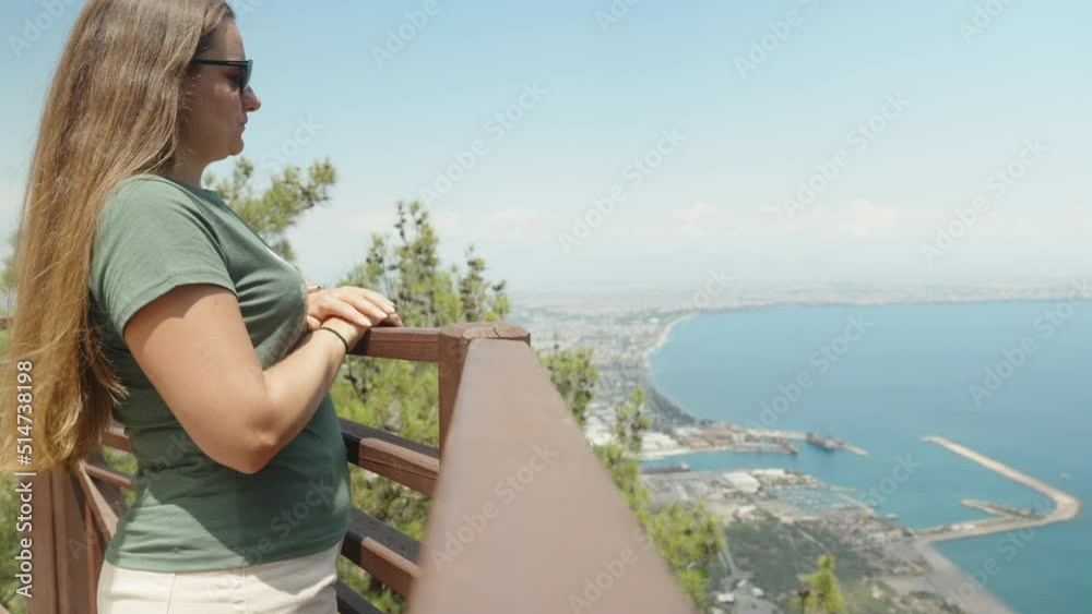 A girl with sunglasses approaches the railing on the top of the mountain. She says something and points to the sea below. Summer walk.