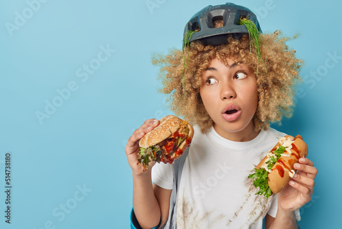 Wondered curly haired young woman eats appetizing hamburger and hot dog wears protective helmet and t shirt smeared with dirt isolated over blue background copy space for your advertisement.