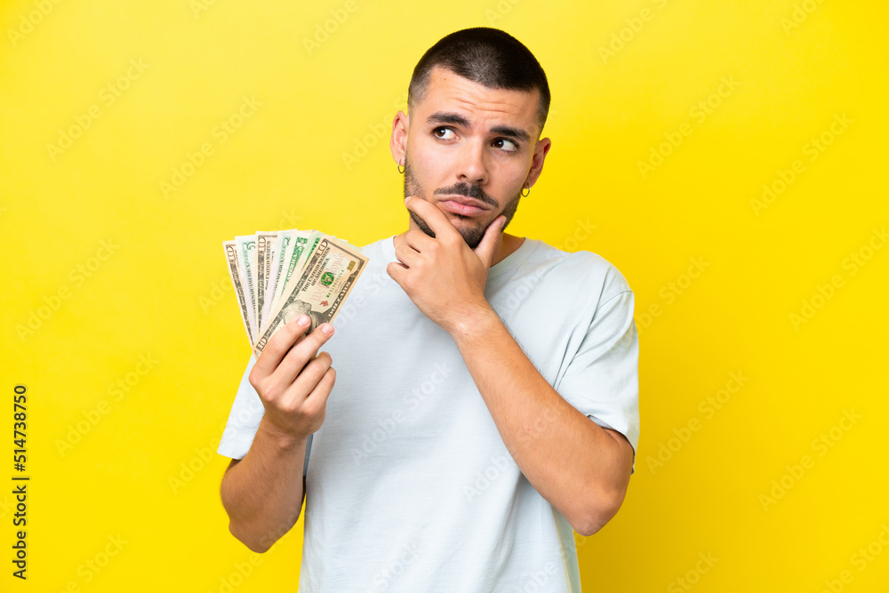 Young caucasian man taking a lot of money isolated on yellow background having doubts Young caucasian man taking a lot of money isolated on yellow background having doubts