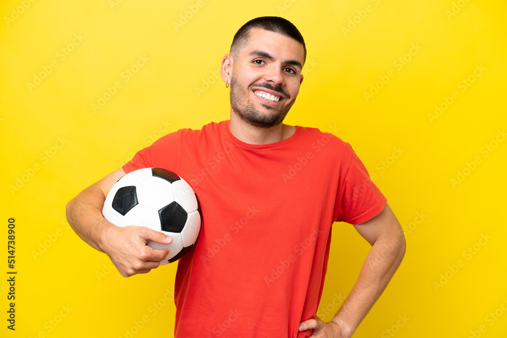 Young caucasian man playing soccer isolated on yellow background posing with arms at hip and smiling