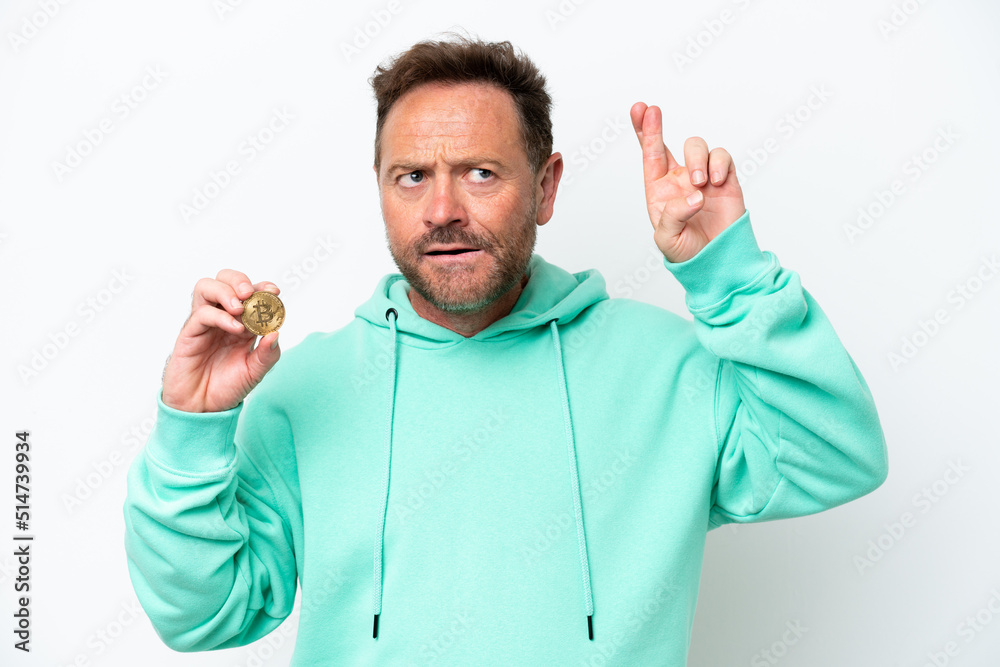 Middle age man holding a Bitcoin over isolated background with fingers crossing and wishing the best Middle age man holding a Bitcoin over isolated background with fingers crossing and wishing the best