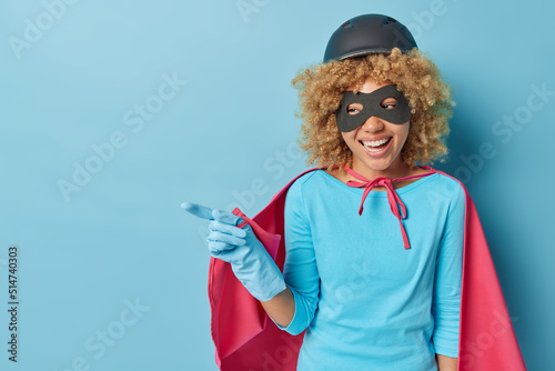 Cheerful female superhero smiles happily points away on blank space prepares for masquerade stands against blue background. Curly haired woman wears mask helmet and cloak shows place for promo