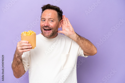 Middle age caucasian man holding fried chips isolated on purple bakcground listening to something by putting hand on the ear © luismolinero