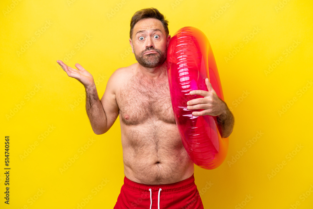 Middle age caucasian man holding inflatable donut isolated on yellow background having doubts while raising hands