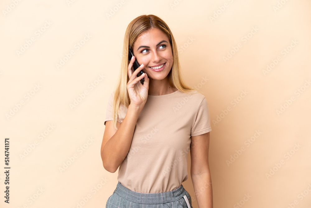 Blonde Uruguayan girl isolated on beige background keeping a conversation with the mobile phone with someone Blonde Uruguayan girl isolated on beige background keeping a conversation with the mobile phone with someone