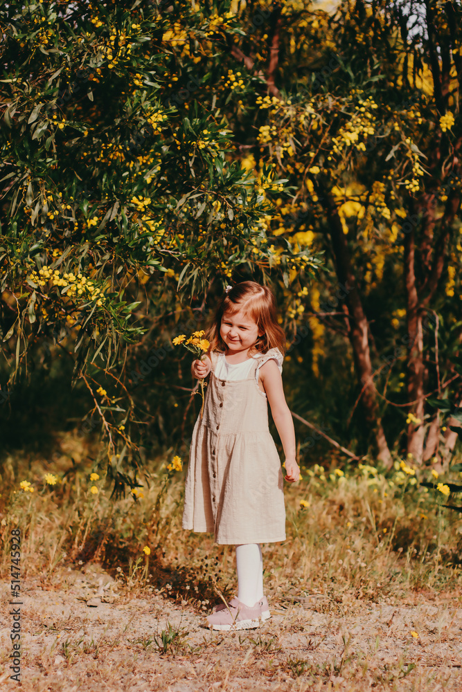 Cute toddler girl with long hairs wear casual dress walking, runing in field with green grass and blooming trees with yellow flowers.