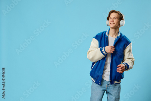a stylish young man in a fashionable jacket and with large white headphones on his head dances to the music while standing against a light blue background. Horizontal photo with plenty of insert space