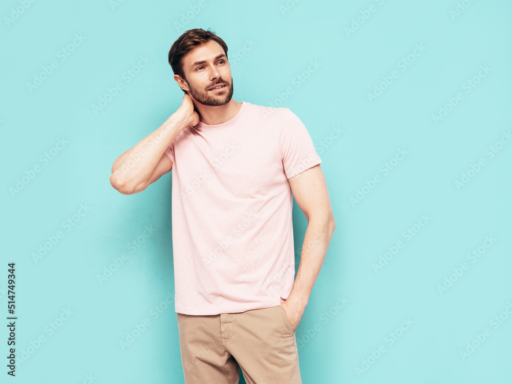 Portrait of handsome smiling stylish hipster lambersexual model. Sexy man dressed in pink T-shirt and trousers. Fashion male isolated on blue wall in studio