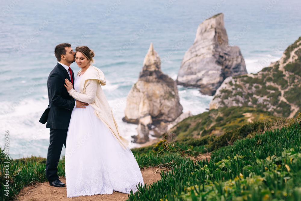 the groom hugs and kisses the bride on ocean coast overlooking t
