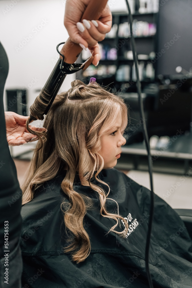 Fototapeta premium Female hands of a hairdresser. Close-up of blond hair on a curling iron. Curling curls. Beauty saloon.