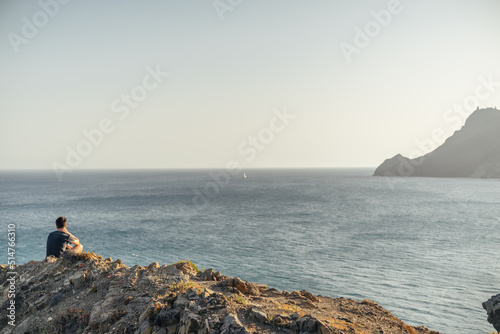 Young man sitting on the edge of a cliff in front of the sea at sunset