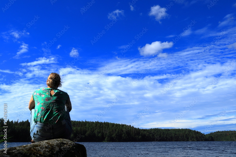 Fototapeta premium Scandinavian summer weather one sunny day. Woman sitting on a rock with back to camera. Next to the Swedish lake Malar or Mälaren. Great view. Järfälla, Stockholm, Sweden, Scandinavia, Europe.