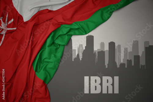 abstract silhouette of the city with text Ibri near waving national flag of oman on a gray background.3D illustration