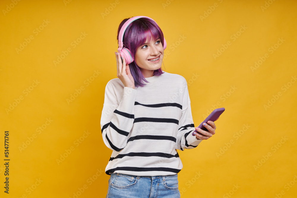 Cheerful young woman with headphones using smart phone while standing against yellow background Cheerful young woman with headphones using smart phone while standing against yellow background
