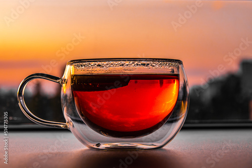 Close up of tea with lemon in transparent cup on table and city at sunset on background. Hot drink with colorful liquid in glass cup with reflections, condensate and mirrored nature, front view.