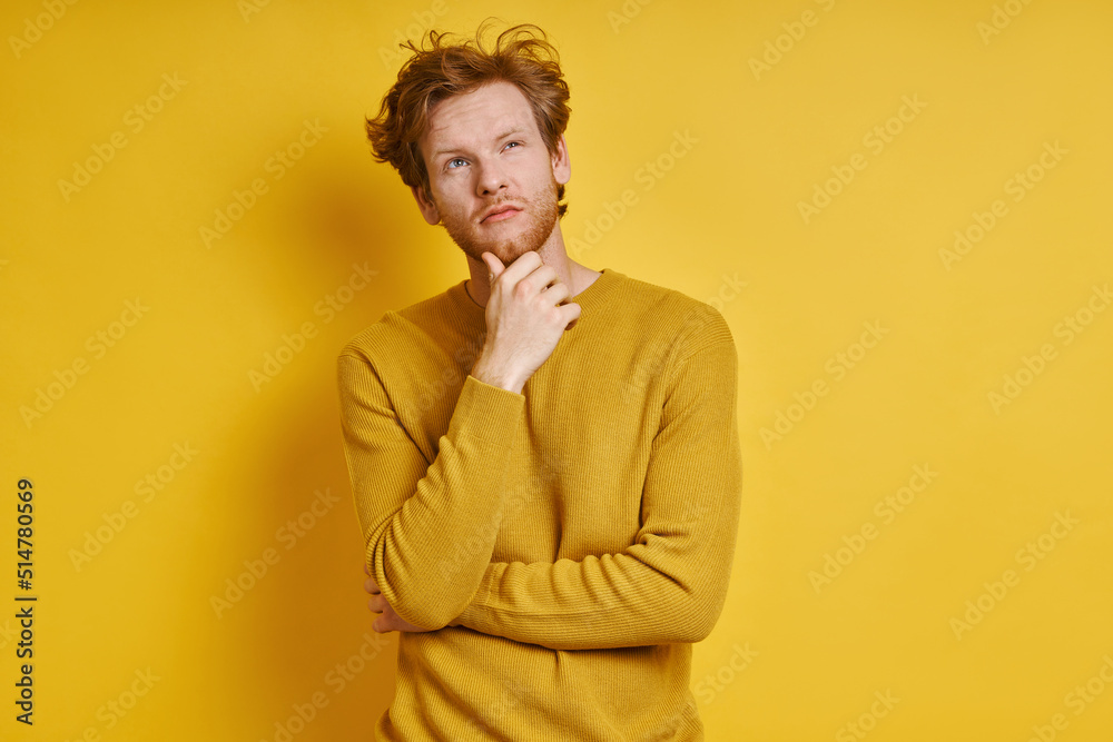 Thoughtful redhead man holding hand on chin while standing against yellow background Thoughtful redhead man holding hand on chin while standing against yellow background