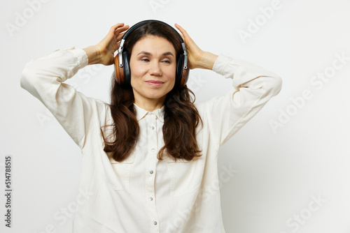 funny woman in light shirt stands listening to music in her headphones, closing her eyes with pleasure and holding headphones with her hands. Horizontal photo on a light background with an empty space