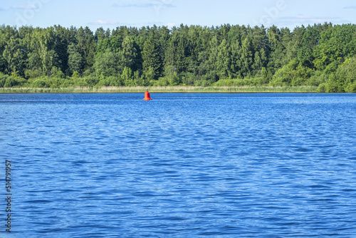 Summer water landscape, blue lake surface and lush green forest foliage on bank. Red buoy in middle of river. Outdoor recreation club, vacation camp,wild nature,copy space. Place for yachting, kiting