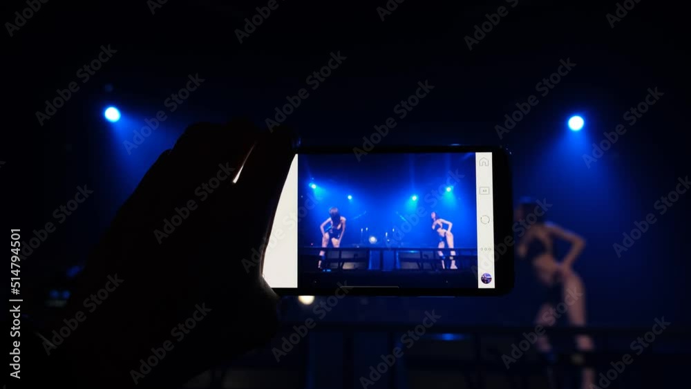 A man's hand holding a smartphone shoots a video of an Asian female model performing a sexy dance on stage.	
