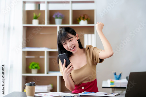 Happy young Asian business woman successful excited raised hands rejoicing with smartphone in the office.