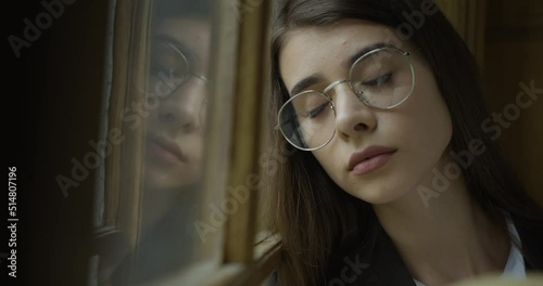 Sad young girl in glasses sits on the windowsill and looks far away photo