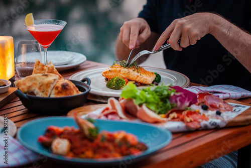 Man on luxury dinner served on table with fresh seafood, cold cuts and cocktail glass in luxury outdoor restaurant. Man hands cutting grilled salmon. Healthy food