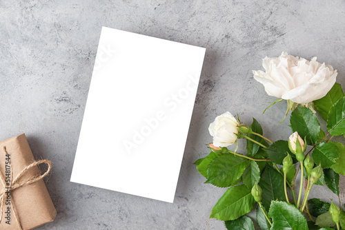 Blank greeting card or wedding invitation with white rose flowers and gift box on gray background. Mock up. Flat lay