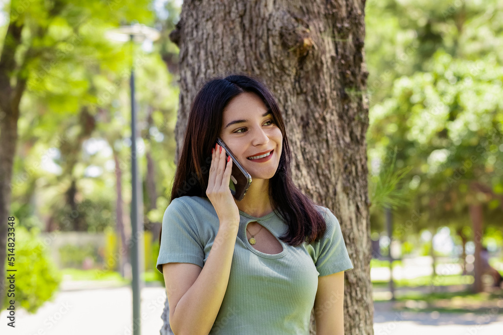 Happy woman wearing tee standing on city park, outdoors talking on mobile phone with friends or boyfriend with smiles. Happy woman wearing tee standing on city park, outdoors talking on mobile phone with friends or boyfriend with smiles.