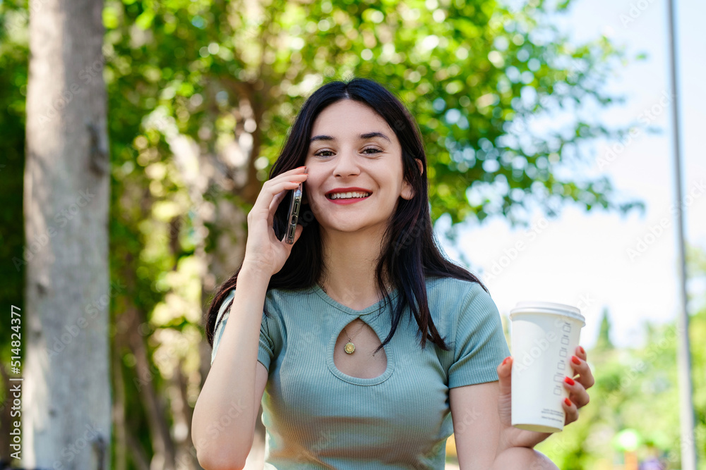 Happy woman wearing tee standing on city park, outdoors holding takeaway coffee mug and talking on phone with friends while looking at the camera. Happy woman wearing tee standing on city park, outdoors holding takeaway coffee mug and talking on phone with friends while looking at the camera.