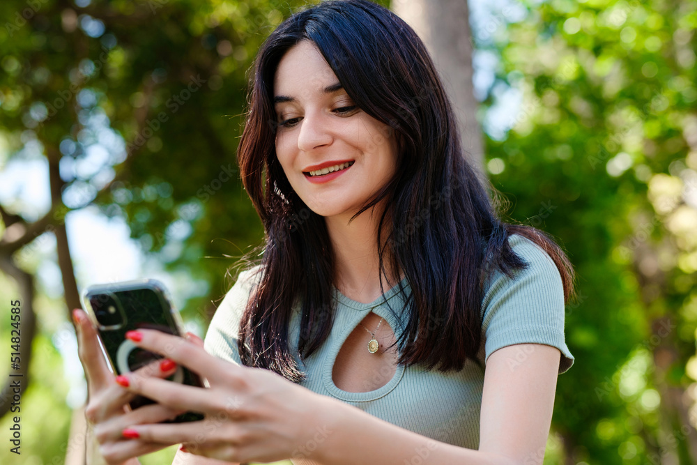 Young brunette woman wearing turquoise tee and orange short on city park, outdoors looking at the phone screen and using phone. Messaging with friends, watching video or scrolling on social media. Young brunette woman wearing turquoise tee and orange short on city park, outdoors looking at the phone screen and using phone. Messaging with friends, watching video or scrolling on social media.