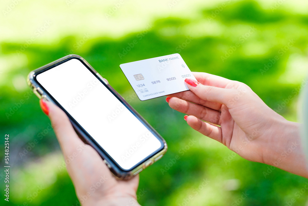 Brunette millennial woman wearing turquoise tee on city park, outdoors holding blank screen mobile phone credit bank card do online shopping provide internet payment. Safe mobile banking.