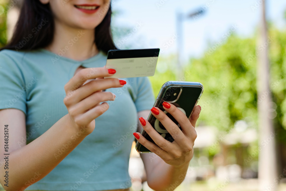 Young woman smiling confident wearing turquoise tee and orange short on city park, outdoors holding mobile phone credit bank card do online shopping provide internet payment. Safe mobile banking. Young woman smiling confident wearing turquoise tee and orange short on city park, outdoors holding mobile phone credit bank card do online shopping provide internet payment. Safe mobile banking.