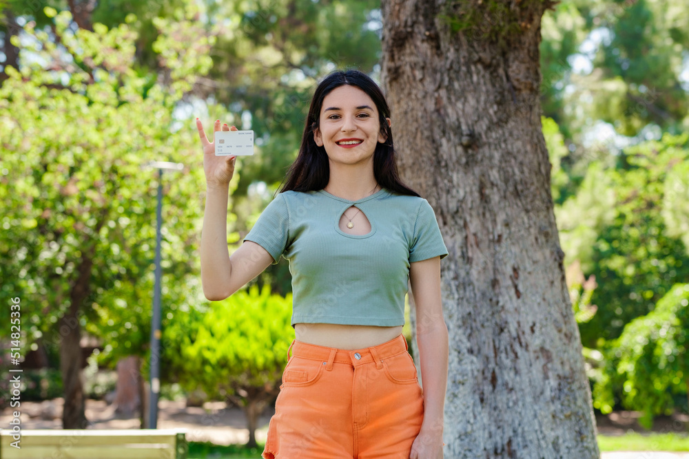 Cute caucasian woman wearing turquoise tee on city park, outdoors showing his plastic credit or debit card with hands. Shopping and finance concept. Cute caucasian woman wearing turquoise tee on city park, outdoors showing his plastic credit or debit card with hands. Shopping and finance concept.