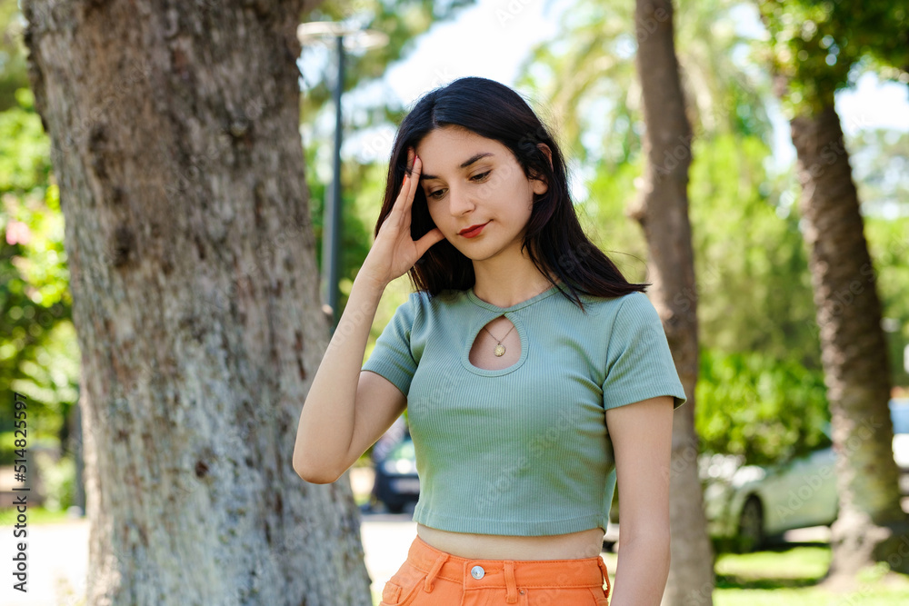 Young brunette woman wearing turquoise tee and orange short on city park, outdoors holding her head with hands trying to remember something or having headache. Suffering from pain.