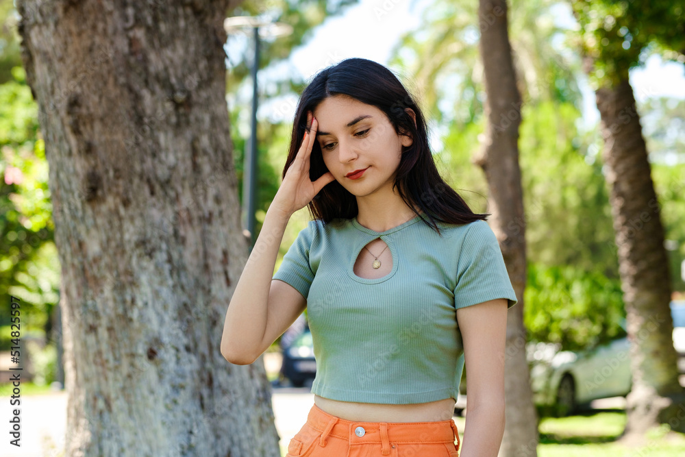 Young brunette woman wearing turquoise tee and orange short on city park, outdoors holding her head with hands trying to remember something or having headache. Suffering from pain. Young brunette woman wearing turquoise tee and orange short on city park, outdoors holding her head with hands trying to remember something or having headache. Suffering from pain.