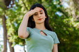 Young caucasian woman wearing tee standing on city park, outdoors holding her head with hands trying to remember something or having headache. Suffering from pain.