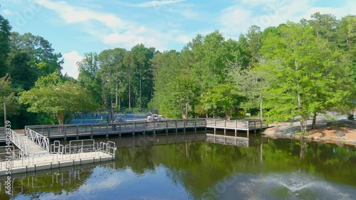 stunning aerial footage of gorgeous summer landscape in the park with a lake and a water fountain and a brown wooden bridge surrounded by pink trees and lush green trees and grass with blue sky photo