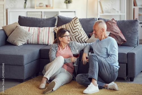 Content relaxed senior couple in casual clothing sitting on floor in living room and drinking red wine while talking