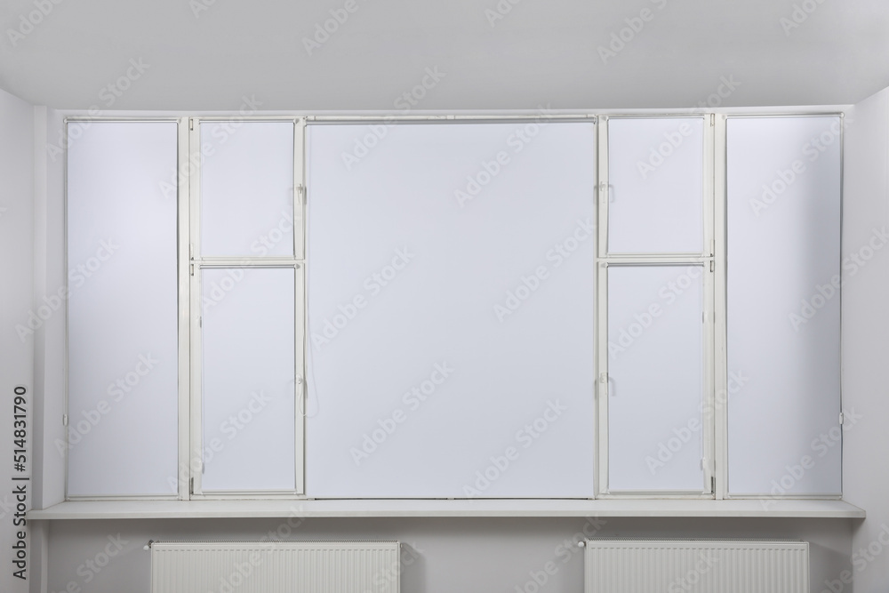 Large window with closed white roller blinds indoors Large window with closed white roller blinds indoors