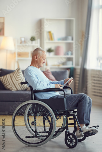 Serious senior man with gray hair sitting in wheelchair and surfing net on tablet while spending time at home