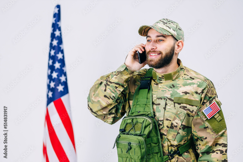 A soldier in camouflage uniform, backpack, happily talking on the phone, receiving good news, standing near the American flag.
